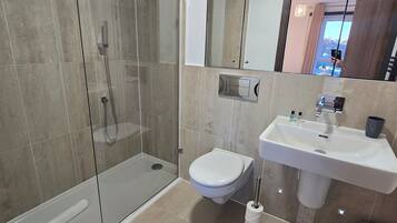 A modern bathroom with a glass-enclosed shower, a white toilet, a sink with a mirror above it, and a trash bin.