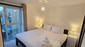 A neatly made bed with white linens, a bedside table with a lamp, and a view of a balcony through a large window. - Tranquil 2 Bed, 2 Bath Apartments walking distance from the Uxbridge town centre (Denham)