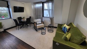 Ein modernes Wohnzimmer mit einem grünen Sofa, einem weißen Sessel, einem Esstisch mit Stühlen und einem an der Wand montierten Fernseher.