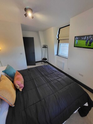 Ein Schlafzimmer mit schwarzer Bettdecke, einer Stehlampe, einem an der Wand montierten Fernseher und einem Fenster mit Jalousien.