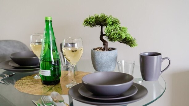 A dining table with a glass top, a green bottle, two glasses of white wine, a bonsai tree, a gray mug, and a bowl set.