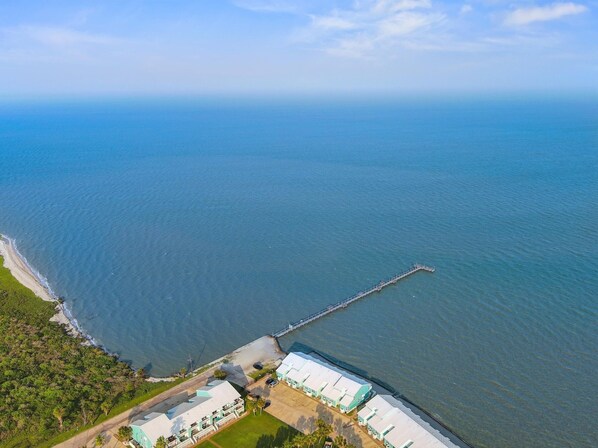 An aerial view of a coastal property with a long pier extending into the sea, surrounded by lush greenery and a series of buildings with white roofs. - Pier Perfection K101 3 Bedroom Condo (Rockport)