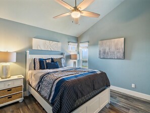 A bedroom with a bed, a nightstand, a lamp, a ceiling fan, and artwork on the walls. - Pier Perfection K101 3 Bedroom Condo (Rockport)