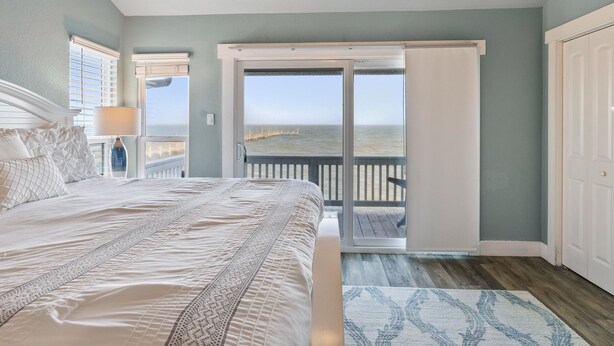A bedroom with a large bed, a ceiling fan, a view of the ocean through a sliding glass door, and a wooden floor. - Pier Perfection K101 3 Bedroom Condo (Rockport)