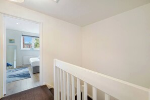 A hallway with a white railing, a door leading to a room with a bed, and a window with a view of buildings. - Stylish 4BR House w/Garden & free parking - ealing (London)