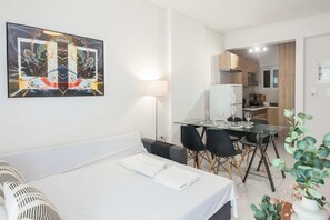A hotel room with a bed, a dining table with chairs, a kitchen area, and a wall-mounted painting. - Olia Urban Luxury Studio (Zografou)