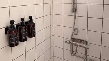 Zona de ducha con cabezal de ducha de mano, ventilador de pared y tres botellas de artículos de aseo.