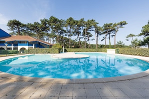 A swimming pool with a tiled deck, surrounded by trees and a building with blue accents. - La Canopée Apartment with Lake View, Shared Pool & Air Conditioning - Conciergerieland (Soustons)