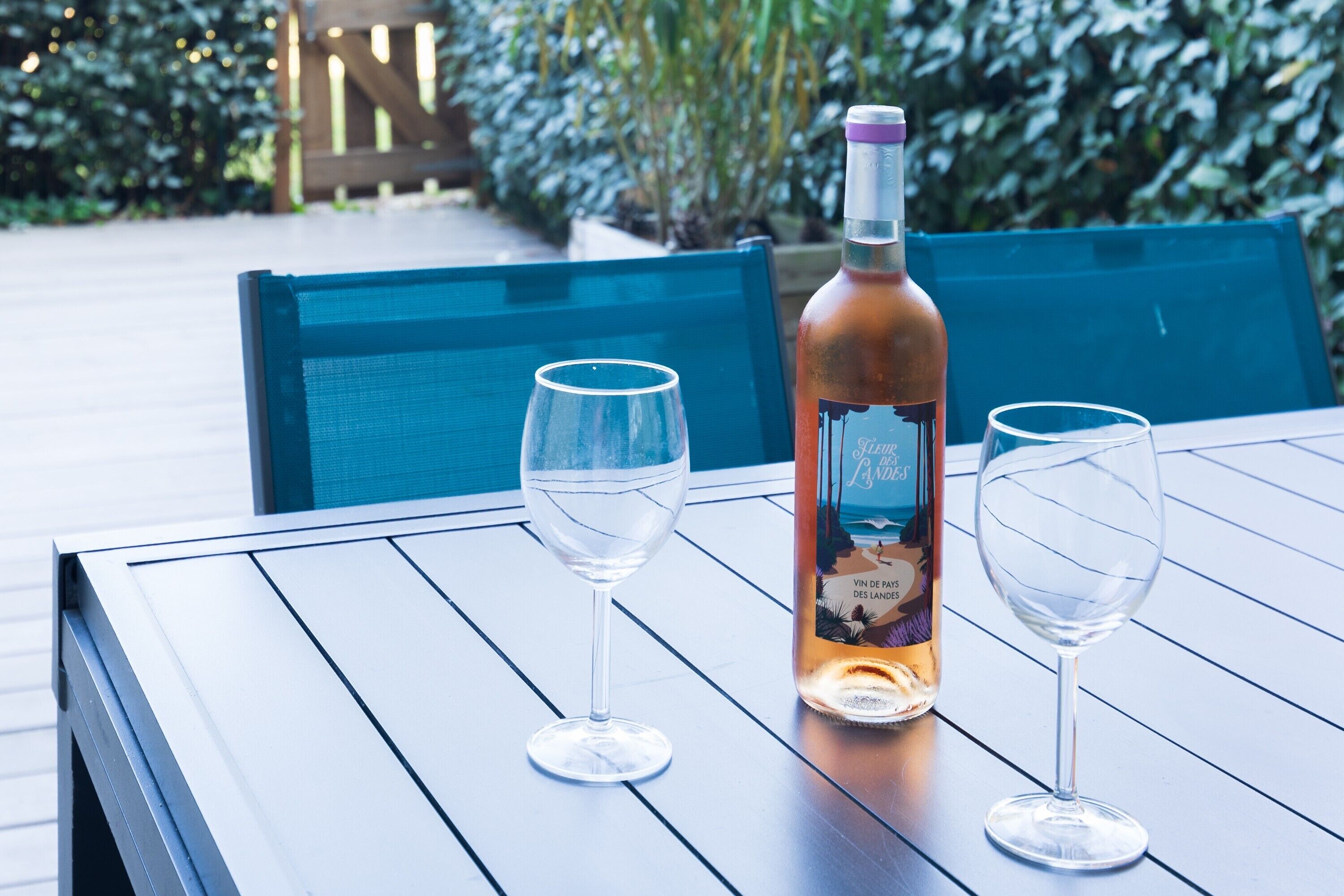 Une table avec une bouteille et deux verres, disposée à l’extérieur, sur une terrasse en bois, avec de la verdure en arrière-plan.
