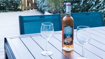 Une table avec une bouteille et deux verres, disposée à l’extérieur, sur une terrasse en bois, avec de la verdure en arrière-plan.