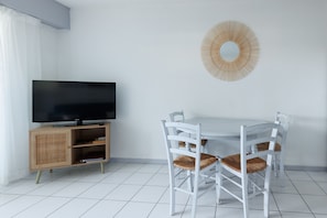 A room with a television on a wooden cabinet, a round wall mirror, a white dining table with four chairs, and a tiled floor. - La Canopée Apartment with Lake View, Shared Pool & Air Conditioning - Conciergerieland (Soustons)
