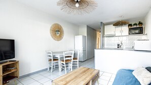 Cozinha moderna com armários brancos, micro-ondas e torradeira. Há uma área de refeições com mesa e cadeiras brancas. Uma TV está sobre um suporte de madeira, e um banco na sala.