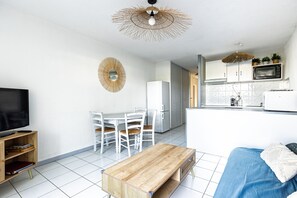 Una cocina moderna con armarios blancos, un microondas y un tostador. Hay una zona de comedor con una mesa y sillas blancas. Un televisor está sobre un mueble de madera, y un banco se encuentra en la sala de estar.