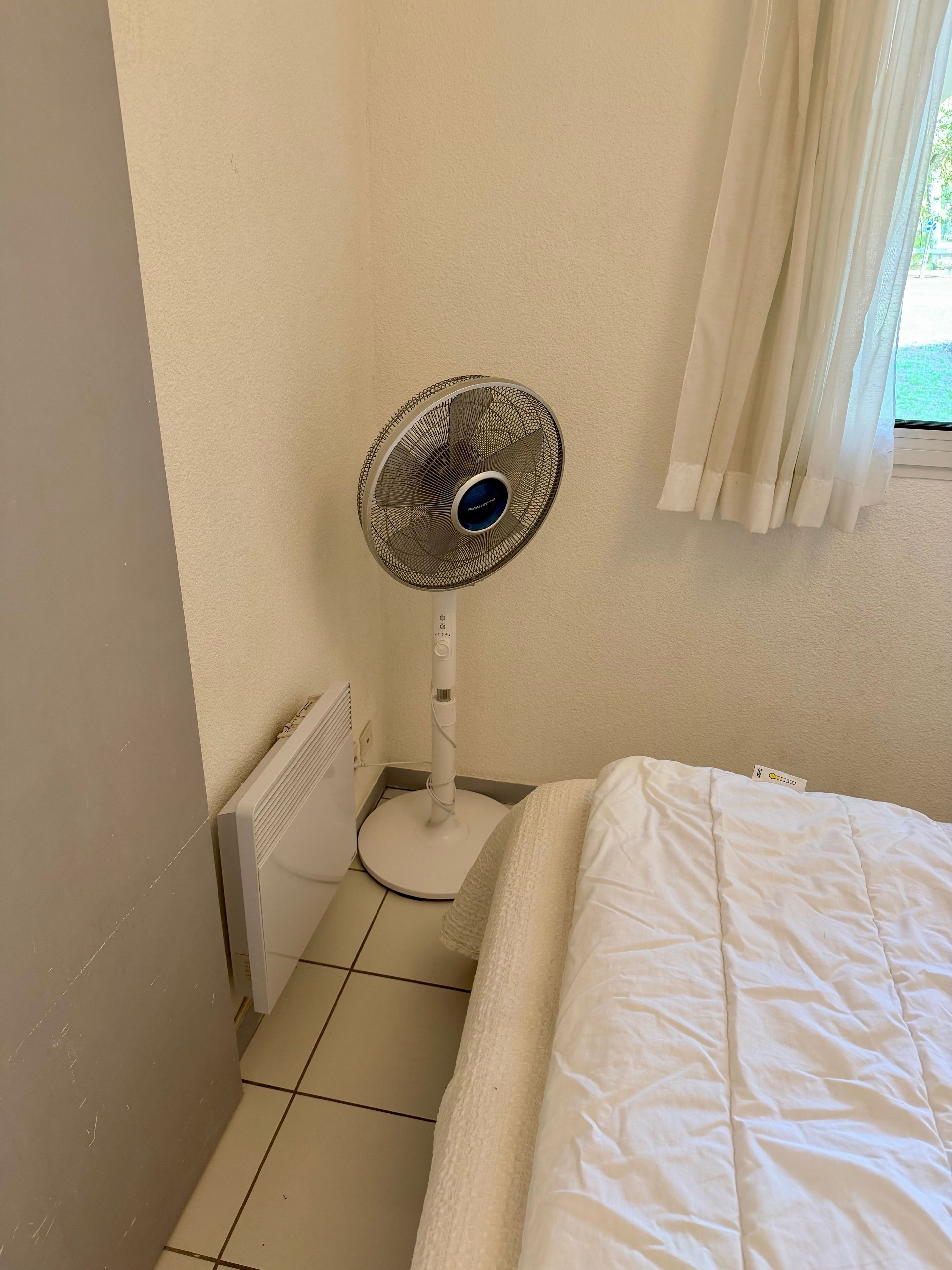 A room with a bed, a standing fan, a radiator, and a window with curtains.