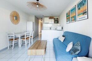 Una sala de estar moderna con un sofá azul, una mesa de centro de madera, una mesa de comedor blanca con sillas y una cocina al fondo.