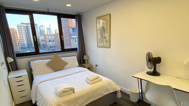 A modern hotel room with a bed, bedside table, lamp, desk, chair, and a city view through the window. - Enjoyable 4-bed, 2.5 bath apartment in the heart of London - Hoxton Zone 1 (London)