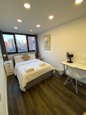 A modern hotel room with a bed, bedside table, lamp, desk, chair, and a city view through the window. - Enjoyable 4-bed, 2.5 bath apartment in the heart of London - Hoxton Zone 1 (London)