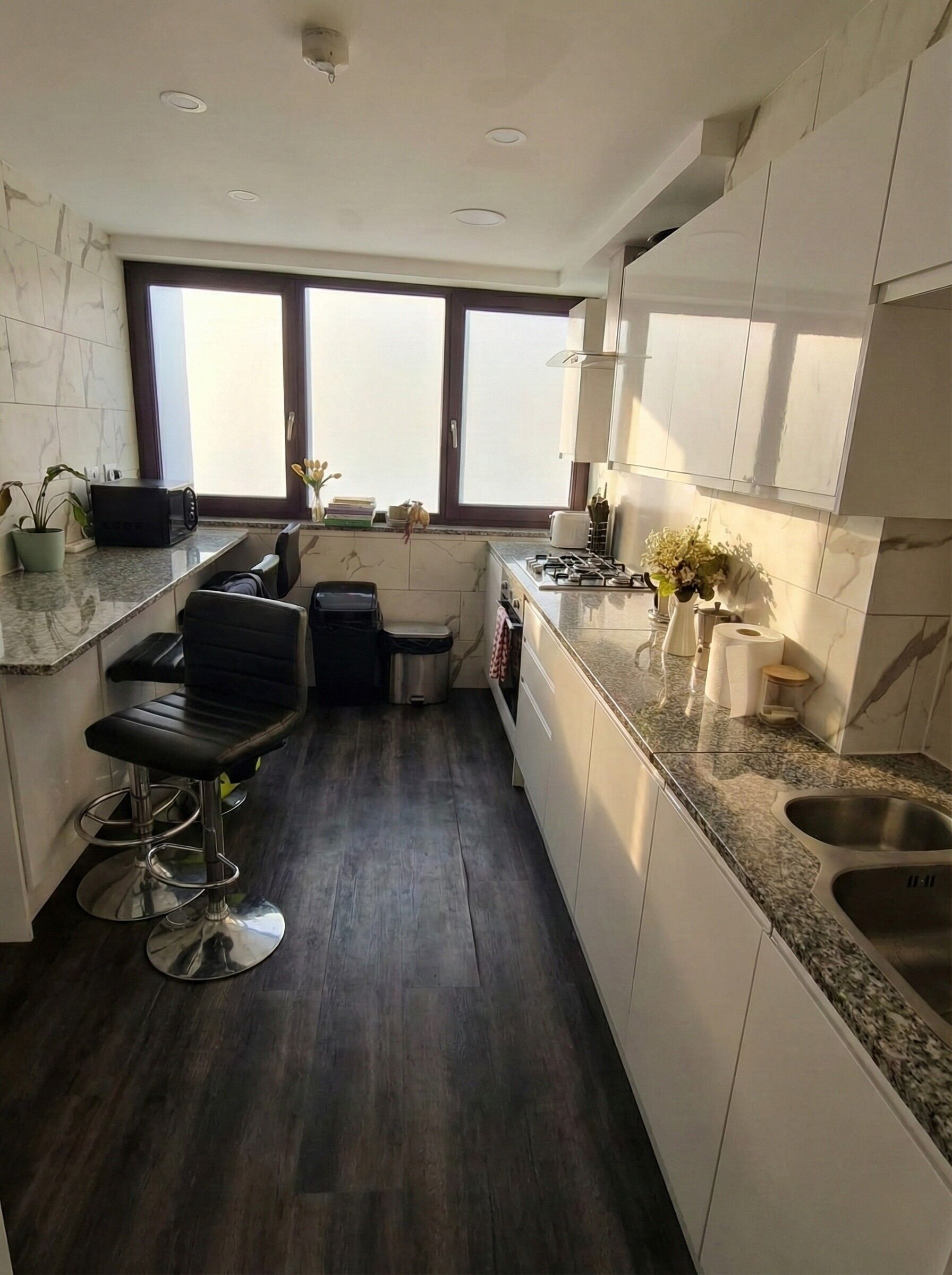 A modern kitchen with dark wood flooring, white cabinetry, a bar stool, and a window providing natural light.
