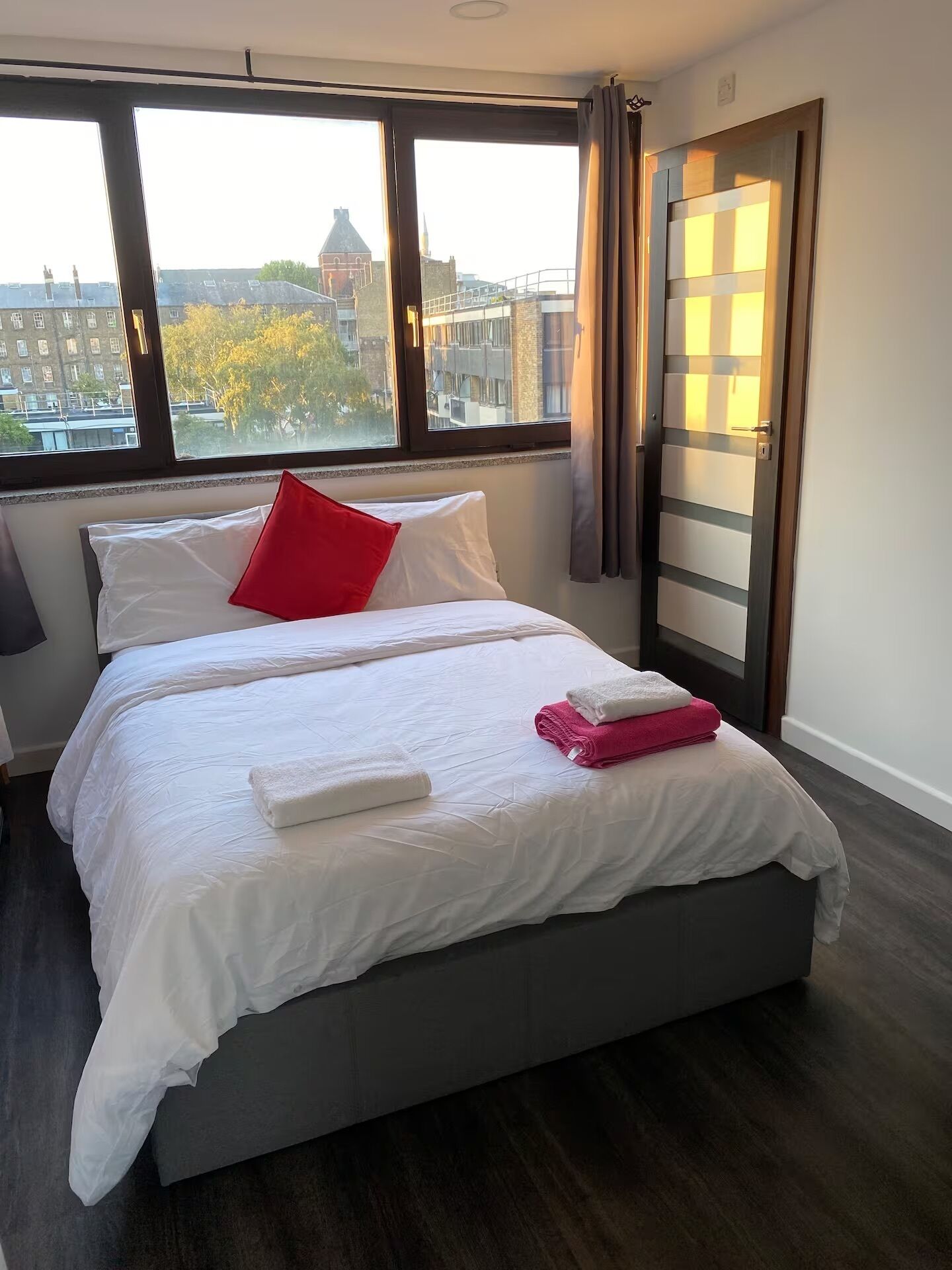 A modern hotel room with a large bed, white bedding, a red pillow, and a view of the city through large windows.