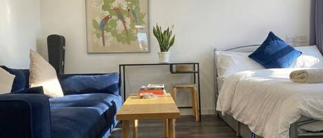 A room with a blue sofa, a bed with white bedding and blue pillows, a small wooden table with books, a potted plant, and a framed picture on the wall.