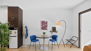 Et moderne køkken med et hvidt køleskab, et mørkebrunt skab og et rundt spisebord med to blå stole. Der er en hængende plante, en gul lampe og et billede i ramme på væggen.