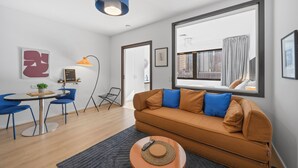 En moderne stue med en brun sofa, blå lænestole, et rundt bord og et stort vindue med udsigt til bygninger.
