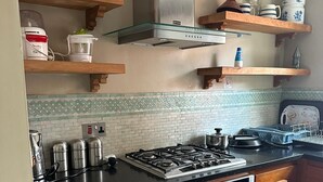A kitchen with a gas stove, oven, and various appliances. There are wooden cabinets, a tiled backsplash, and shelves with kitchenware. - Where English Charm Meets Moroccan Soul - 1 Minute from Kensal Green Station (London)