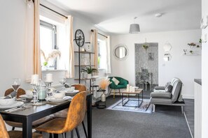 Een moderne eetruimte met een tafel voor vier personen, een groene fauteuil en een grijze bank in een lichte woonkamer met een groot raam.