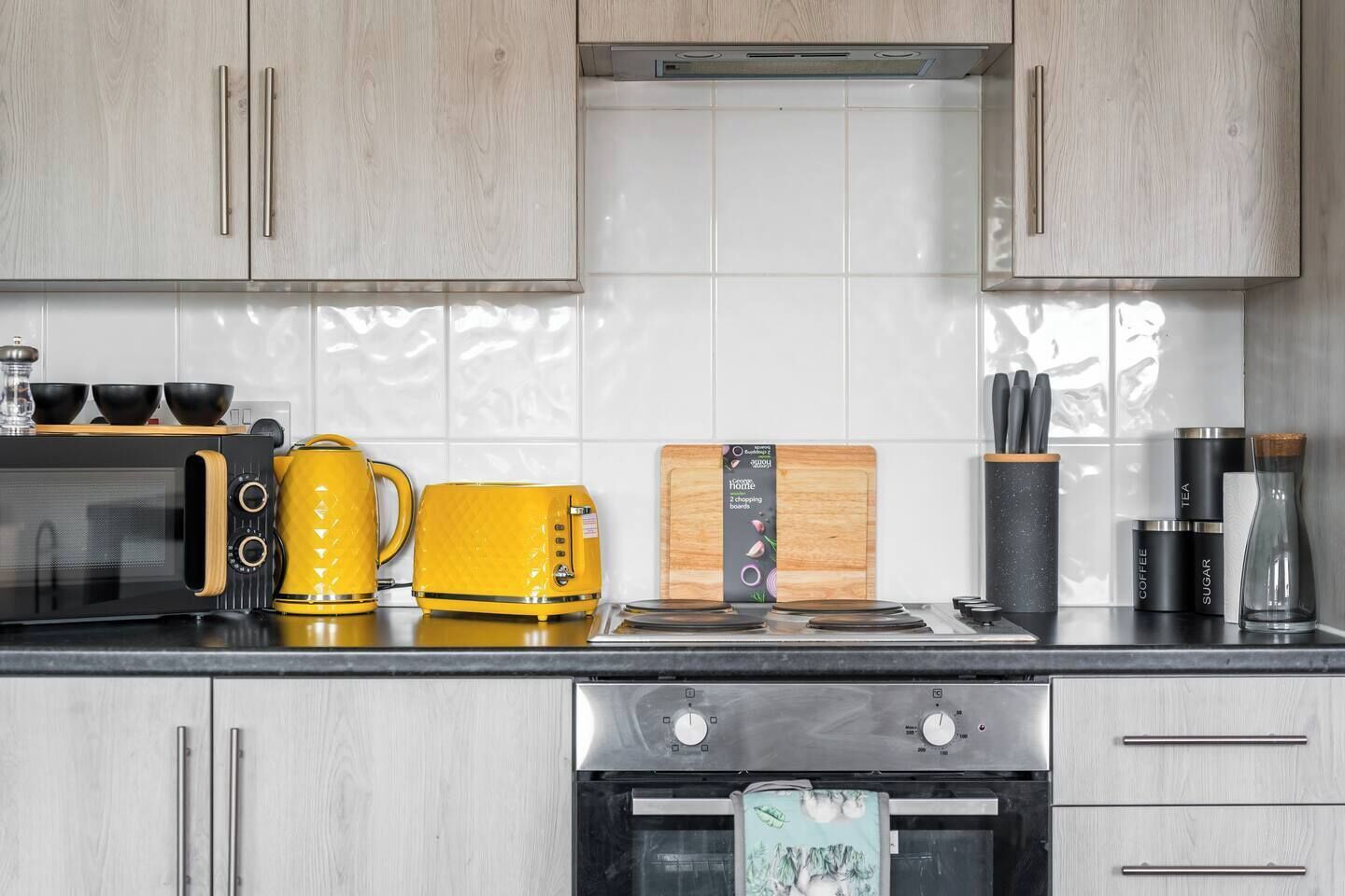 A kitchen with light wood cabinets, a black microwave, a yellow kettle, a cutting board, and a coffee maker.