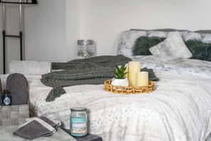 A neatly arranged bedroom with a bed, pillows, a bedside table with a candle, a jar of essential oils, and a basket with candles and a plant. - Modern 1-Bed Apartment with Parking near Heathrow (Slough)
