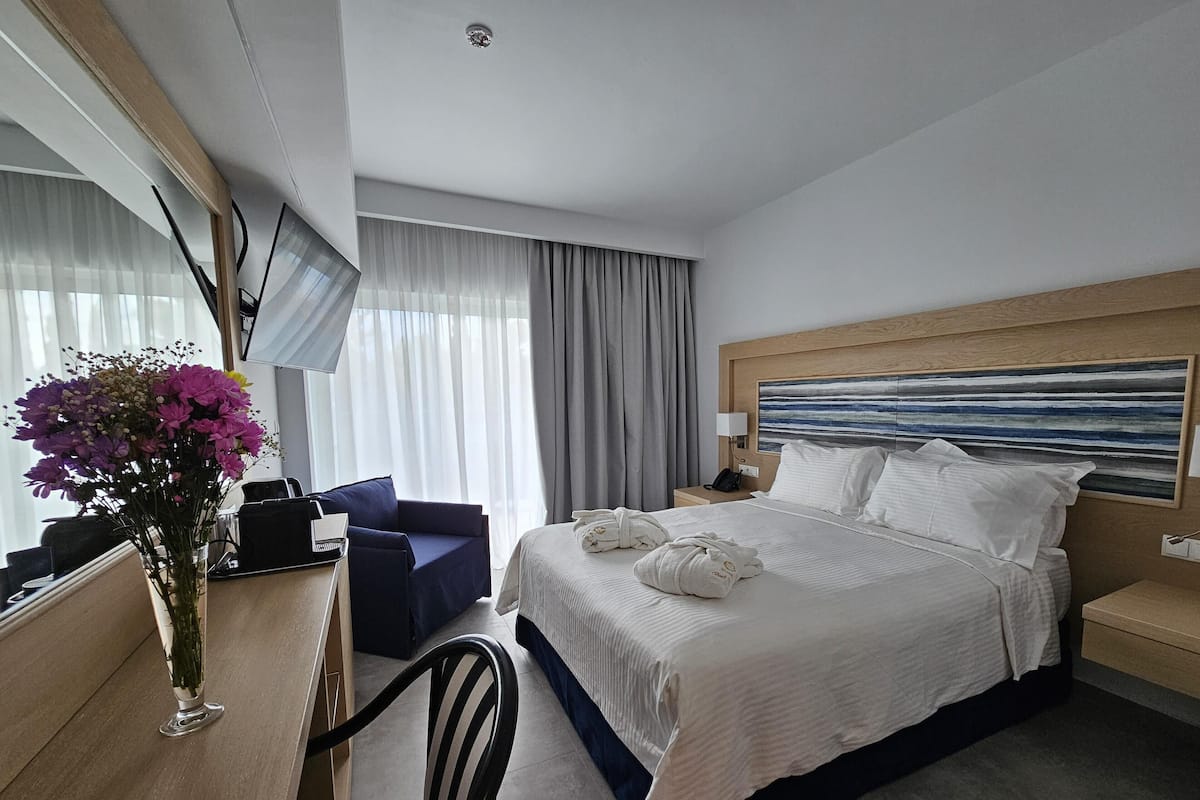 A hotel room with a bed, a desk with a chair, a vase of flowers, a lamp, and a painting on the wall.