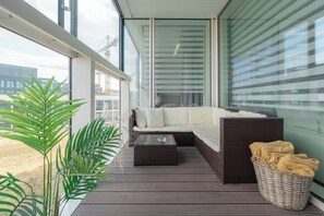 Ein Balkon mit einem Korbsessel, einem Glastisch, einem Korbkorb mit Handtüchern und einem großen Topfpflanzen.