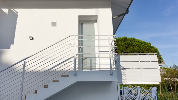 A modern building with a white exterior, a glass door, and a metal staircase leading to a balcony with greenery. - Apartment 'Teresa' with Shared Pool, Private Terrace and Air Conditioning (Bibione)