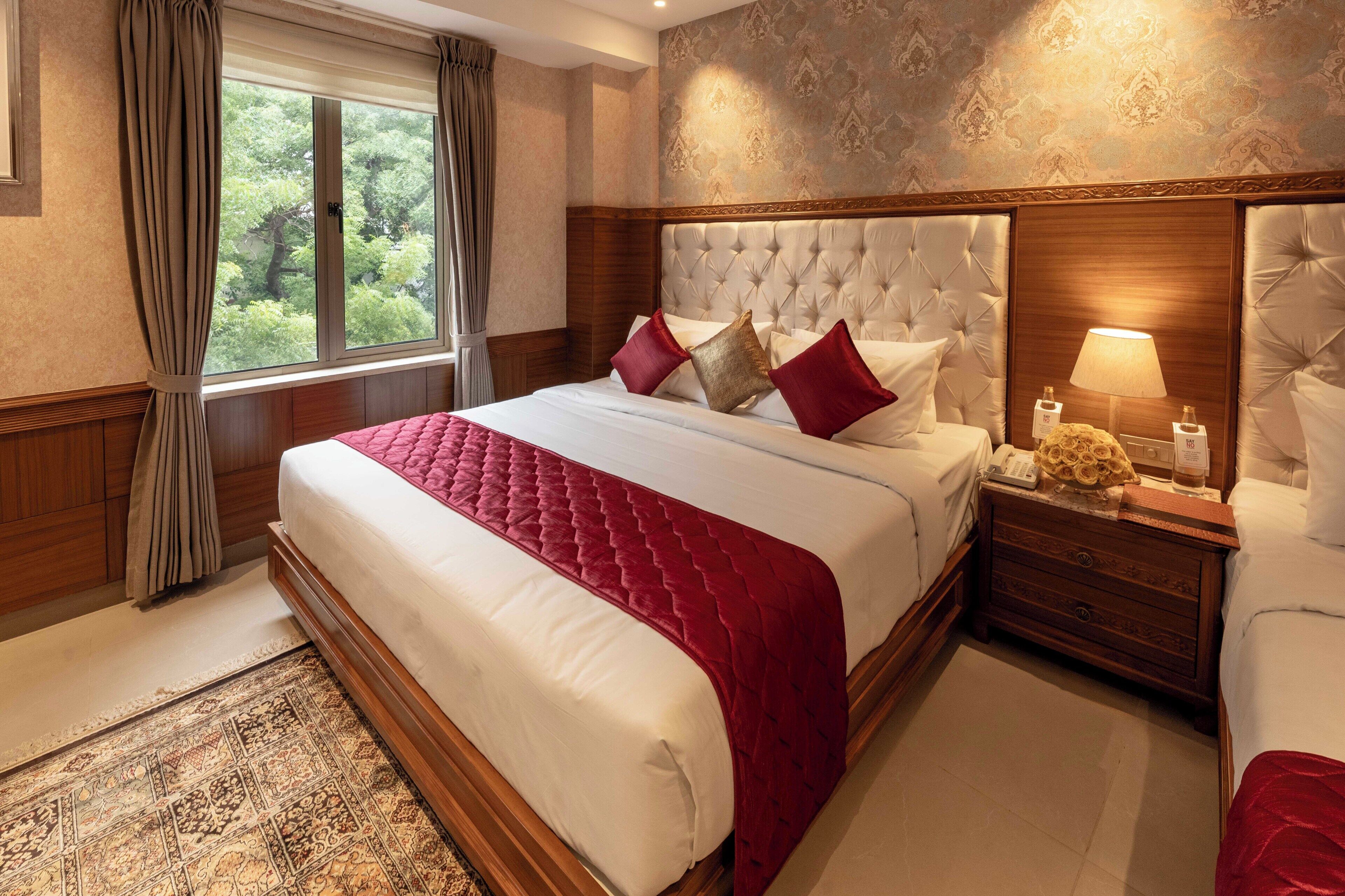 A hotel room with a large bed, a nightstand, a lamp, and a view of trees through the window.