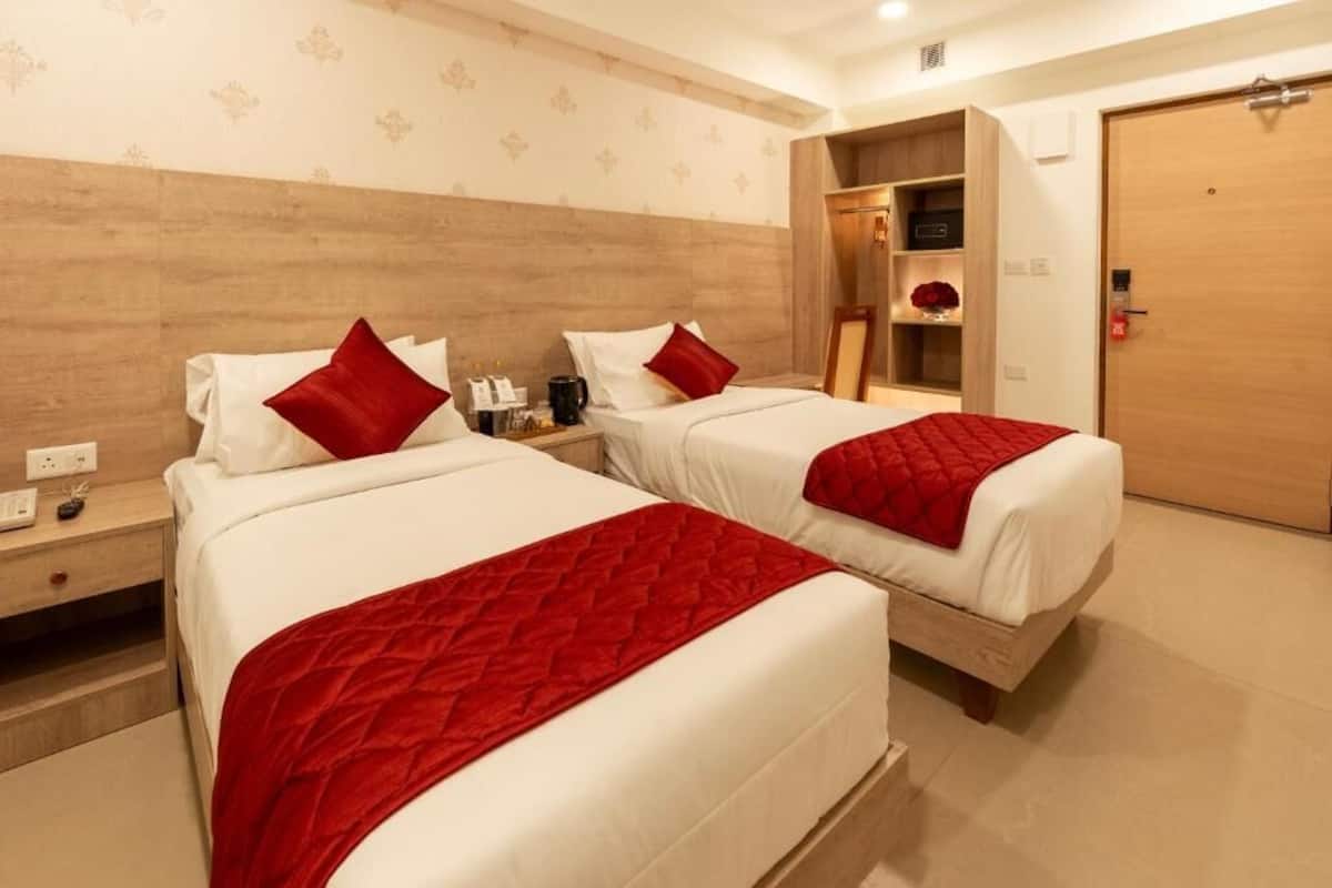 A hotel room with two beds, each with white bedding and red pillows. There is a wooden headboard, a bedside table with a lamp, and a wardrobe.