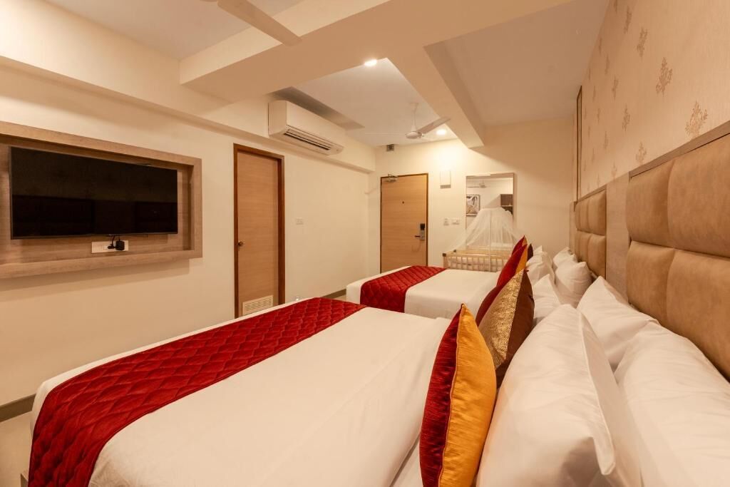 A hotel room with two beds, each with a red and white bedspread, a wooden headboard, and a large flat-screen TV mounted on the wall.