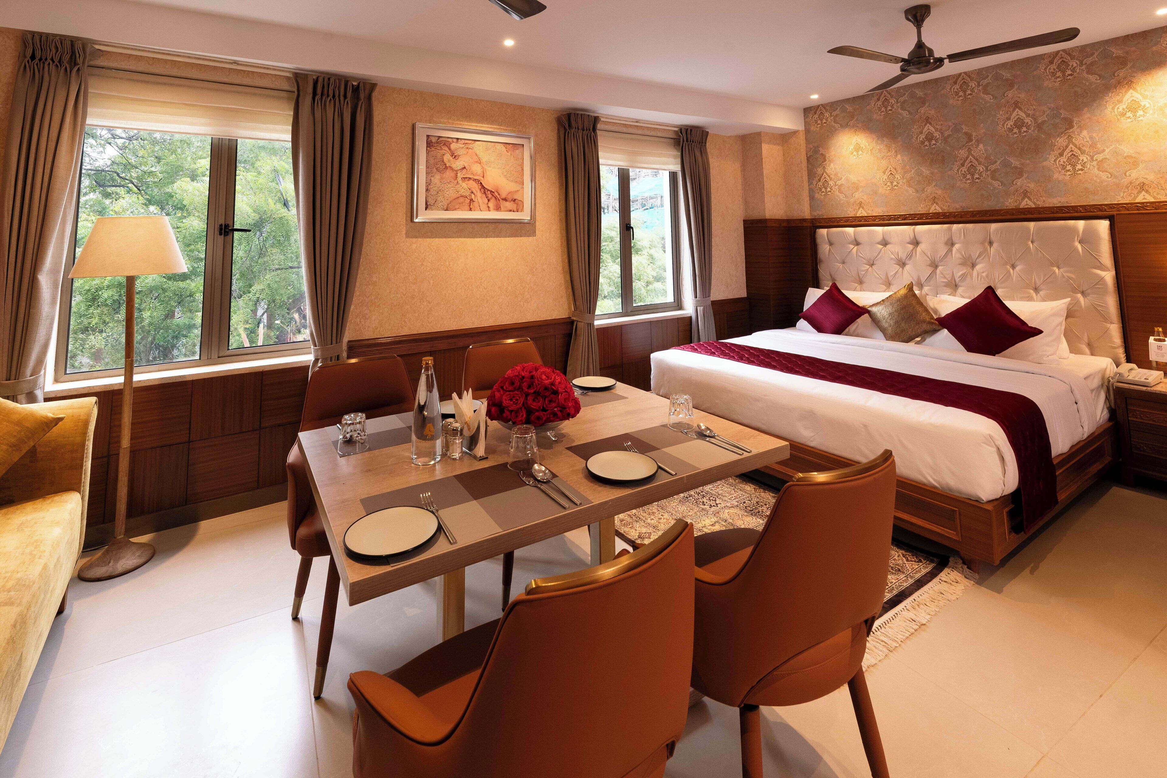 A hotel room with a large bed, a dining table with chairs, a ceiling fan, and large windows with curtains.