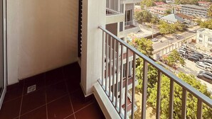 Un balcón con barandilla blanca, un equipo de aire acondicionado y vista a un paisaje urbano.