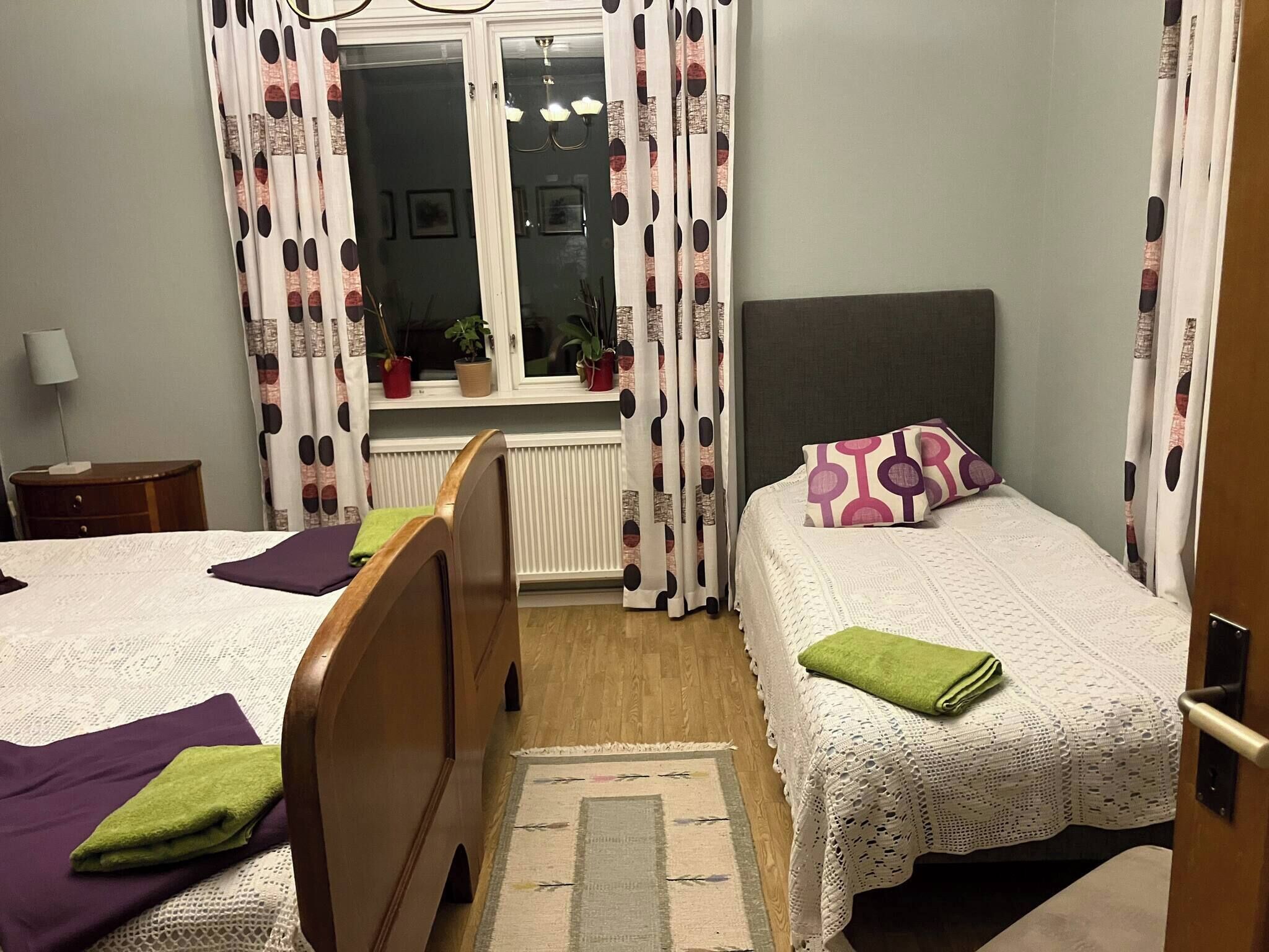 Ein Schlafzimmer mit zwei Betten, einem Heizkörper, einem Fenster mit Vorhängen und einem Nachttisch.