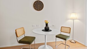 Ein Zimmer mit einem runden weißen Tisch, zwei Korbstühlen mit grünen Kissen, einer Wanduhr, einer Stehlampe und einer Vase mit Blumen.