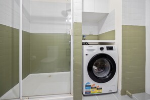 A compact bathroom with a glass-enclosed shower, a white washing machine, and green-tiled walls. - Central Emporium | Urban Oasis 1BR w/King Bed 4max (Melbourne)