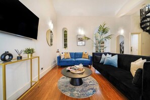 Un salon avec un canapé bleu, un canapé noir, une table basse ronde et un téléviseur fixé au mur.