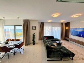A modern living room with a sofa, a dining table with chairs, a TV mounted on the wall, and large windows with curtains.