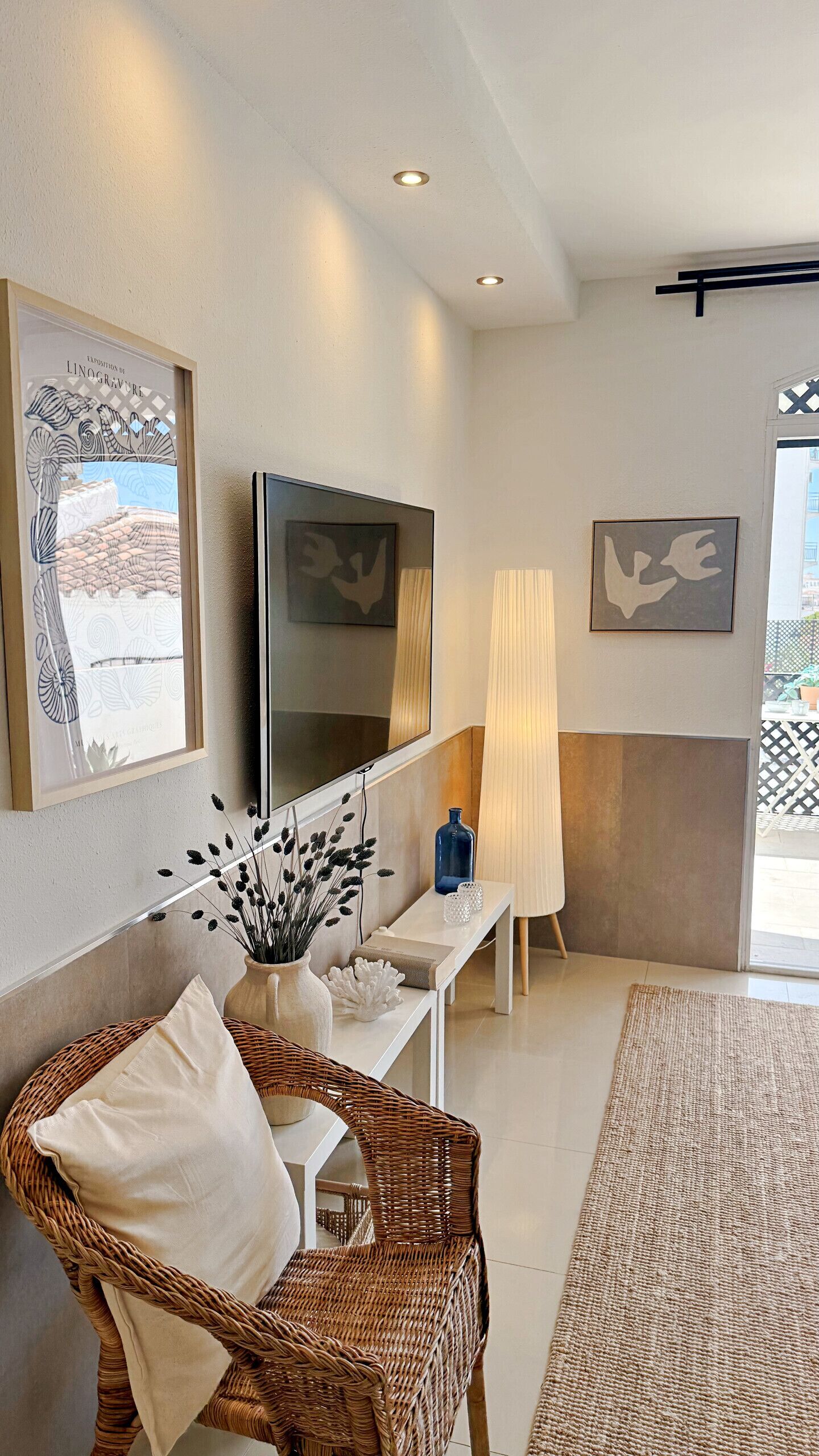 A modern living room with a large flat-screen TV, a wicker chair, a white console table, and artwork on the walls.