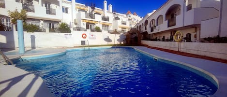 A swimming pool surrounded by white buildings with balconies and a clear blue sky.