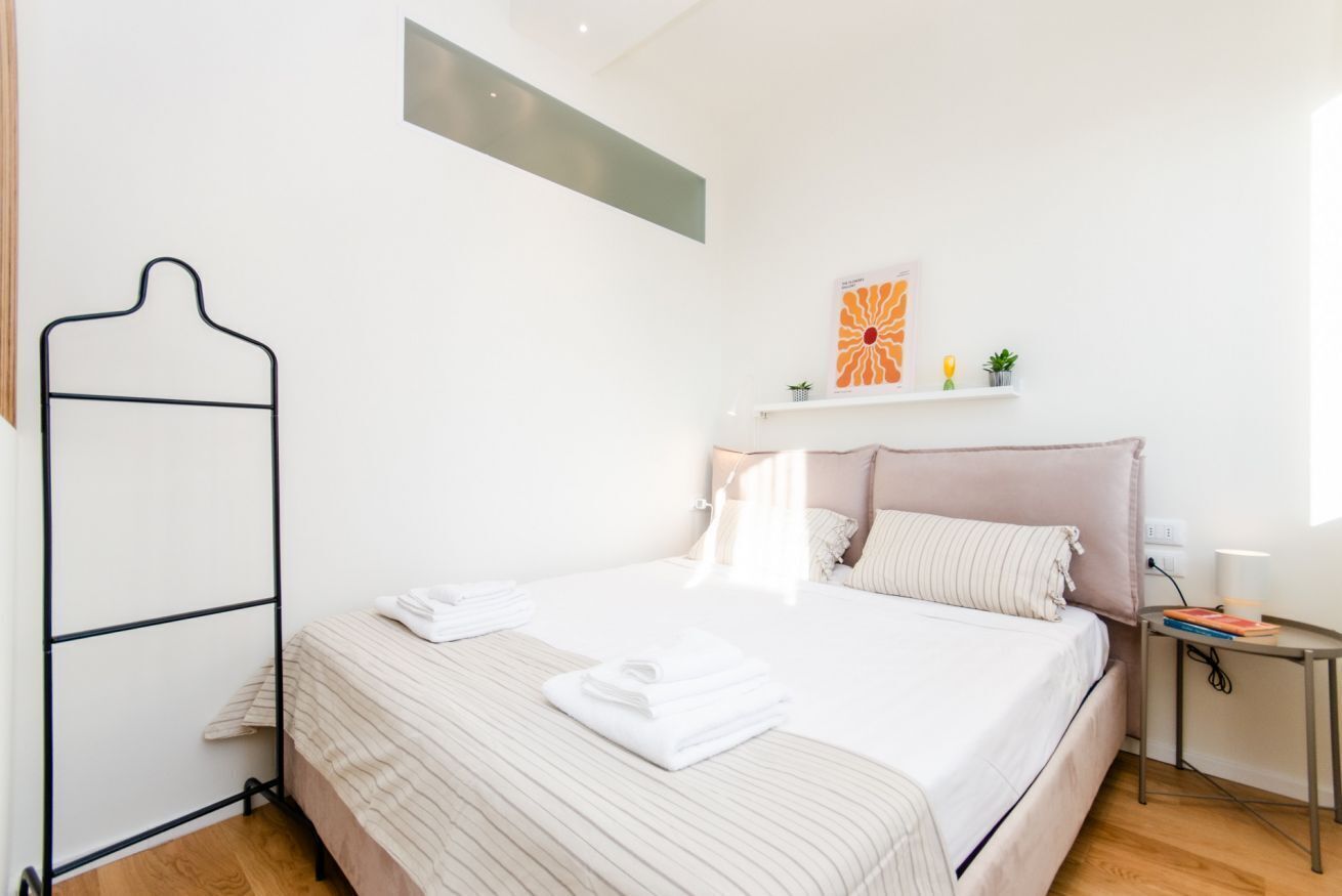 Un letto rifatto con lenzuola bianche e biancheria a righe, un comodino con una lampada e una mensola a muro con oggetti decorativi.