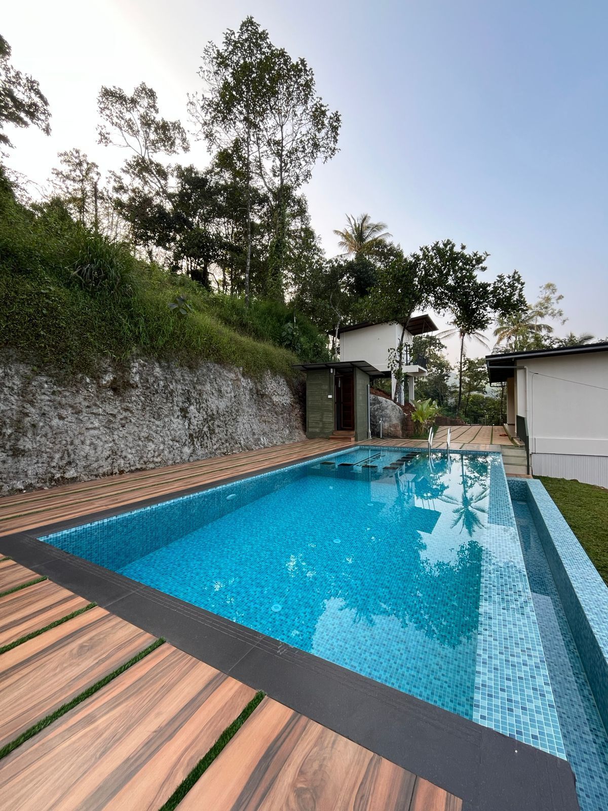 Una piscina rectangular con borde de azulejos, rodeada por una cubierta de madera y un muro de piedra.