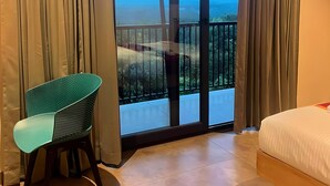 Une chambre avec un lit, une chaise et un balcon offrant une vue sur la verdure et un paysage lointain.