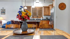 A dining area with a round table, chairs, a vase with flowers, wooden cabinets, and a kitchen in the background. - Moose Mgmt 1206 - Sleeps 7 - Corner with views (Park City)