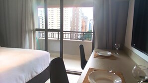 Camera d'albergo con un letto, un tavolo da pranzo apparecchiato con piatti e bicchieri da vino, una sedia e vista sugli edifici attraverso ampie finestre.
